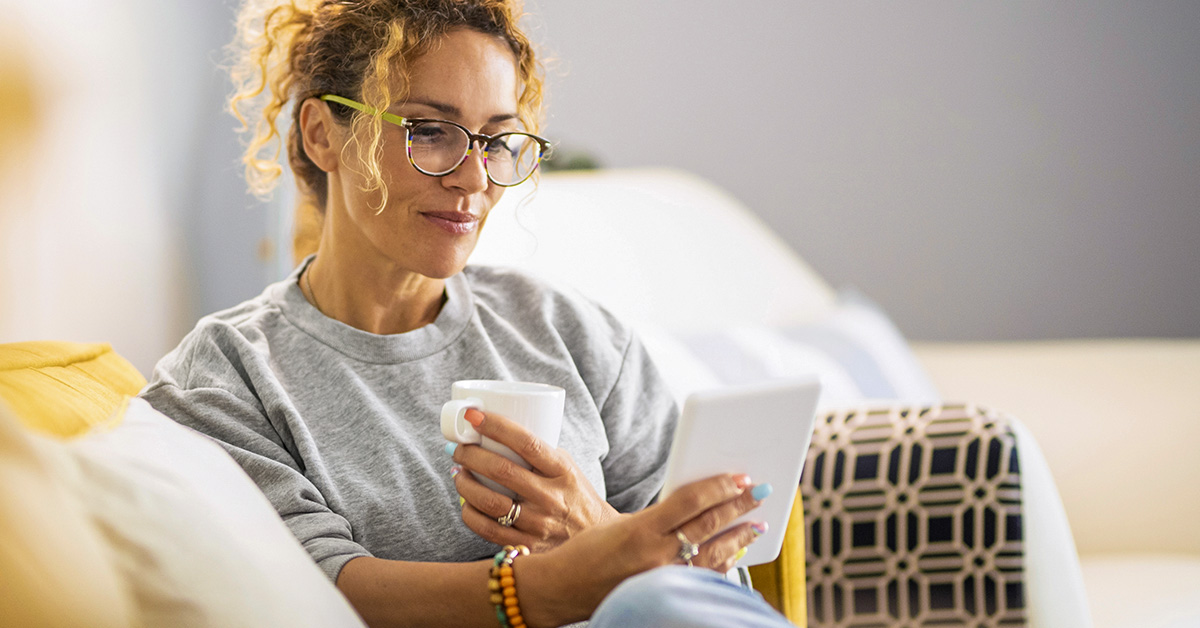 A woman sitting on a couch holding a white mug and looking at budget management tips on a tablet.