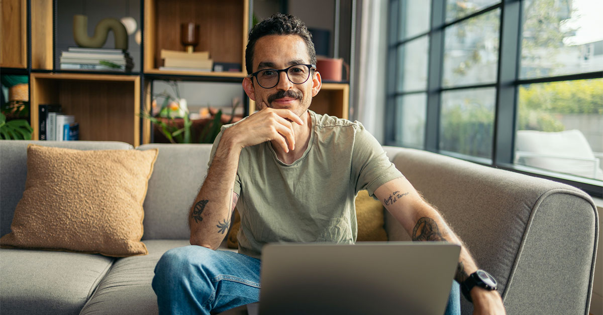  A man sitting on his couch in front of a laptop, smiling at the camera as he manages his personal finances.