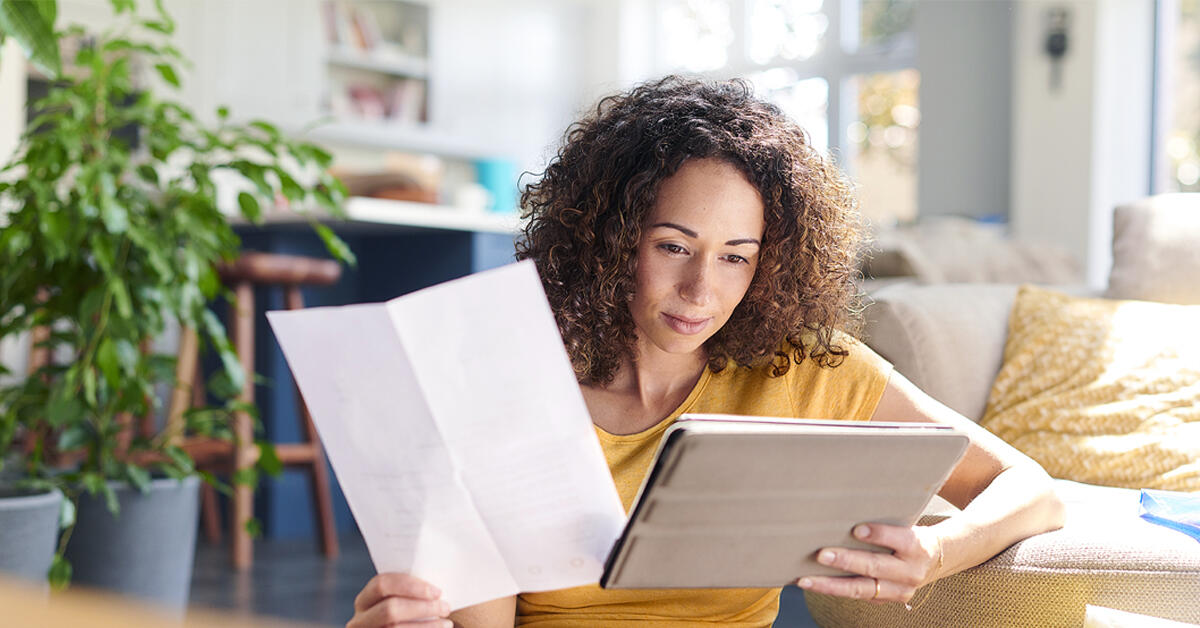 A woman is in her living room holding a paper in her right hand and studying the tablet she is holding in her left hand.