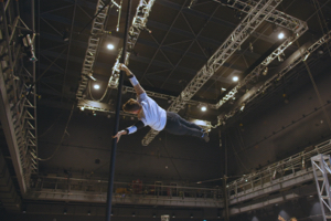 An acrobat swinging around a vertical pole by his hands, legs outstretched