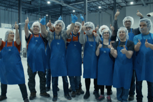A group of employees wearing hair nets, gloves and blue aprons, each giving two thumbs up